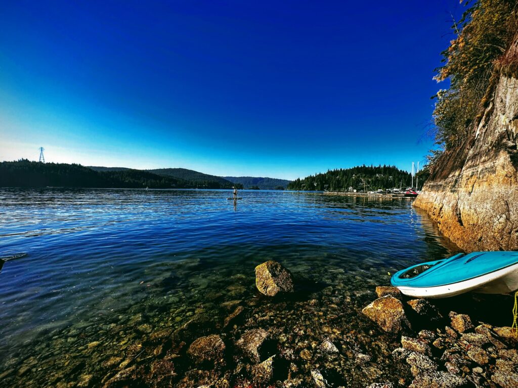 A vibrant blue kayak on the rocks with a paddleboarder on the water in Vancouver, representing Upcycled Unity wellness.